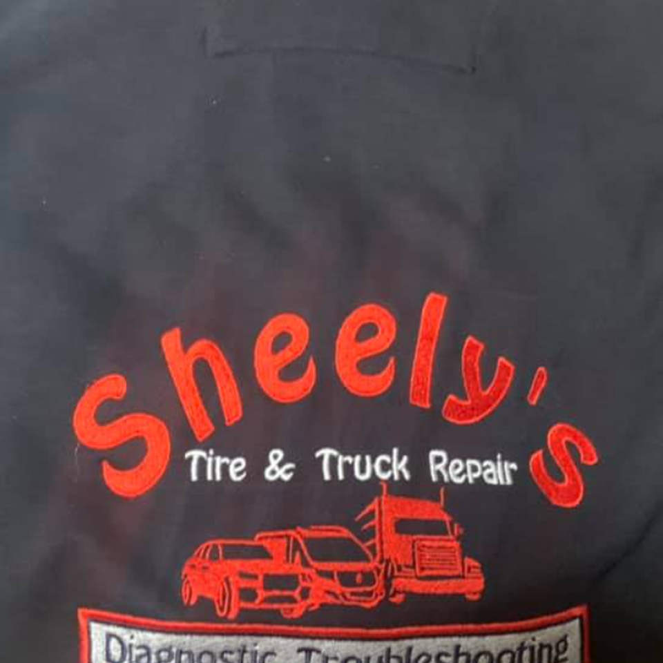 Black embroidered shirt displaying Sneely’s Tire & Truck Repair logo with car and truck graphics and diagnostic troubleshooting text.