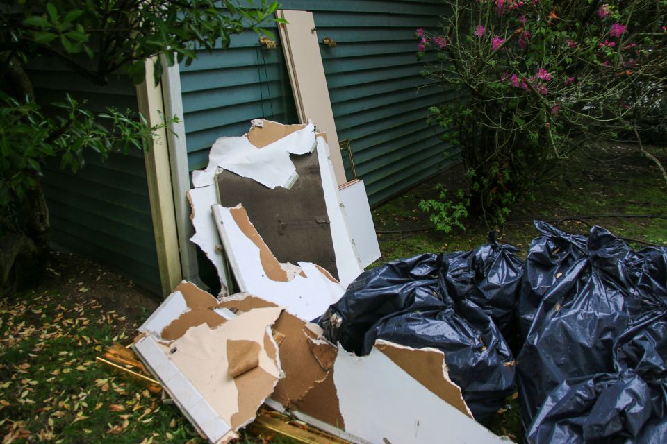 Broken pieces of wall and black bags are discarded near a green structure outdoors