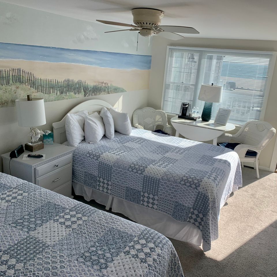 hotel room with a seascape mural on the wall.  2 queen sized bed with light blue patchwork quilt.  Window looking out to a parking lot and craigville beachh