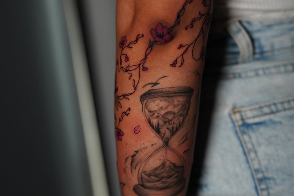 Image of a woman's forearm with an hour glass tattoo in black and white ink and surrounded by ivy in with red flowers