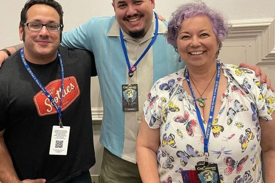 Three panelists smiling together at Con-Cruders 2023, wearing lanyards and badges