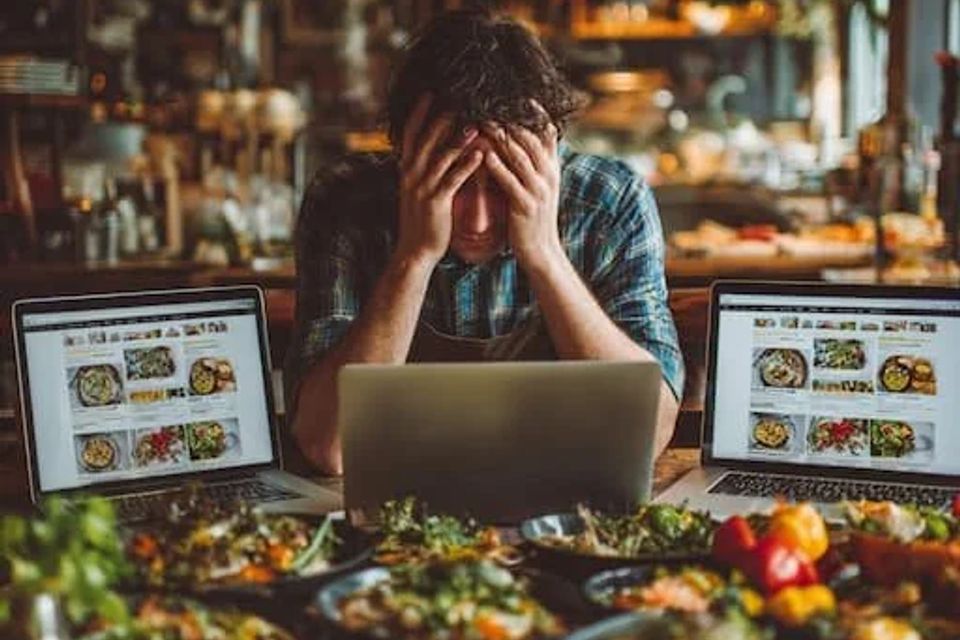 Frustrated restaurant worker sitting around a table full of food and three laptops 