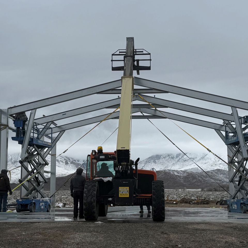 Telehandler lifting steel roof framing during metal building construction with snowy mountains behind