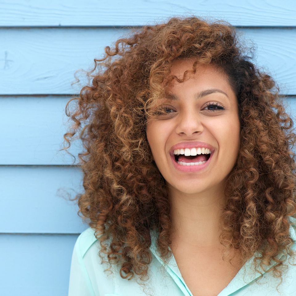 Cheerful young woman with curly hair smiling against a blue wall after Alleviate vitamin IV therapy