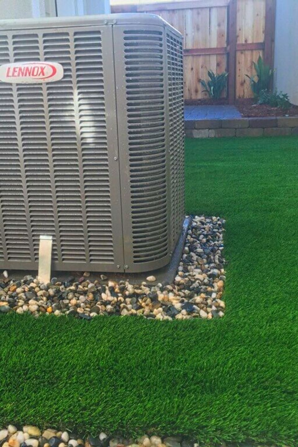 Professional artificial turf installation in a side yard around a Lennox HVAC unit, featuring a clean rock border for drainage and low-maintenance landscaping
