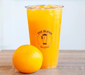 A clear cup of orange smoothie with ice, branded "The Blend Smoothies," placed beside a whole orange on a wooden surface.