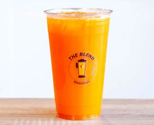 A clear cup of bright orange juice labeled "The Blend Smoothies," placed beside a whole orange, representing a fresh carrot and orange juice blend.