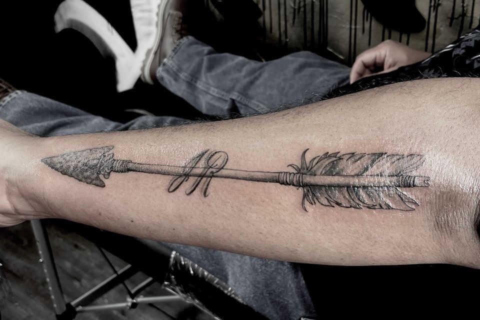 Image of a man's outer calf tattoo of a black and grey arrow with a hand made arrowhead and feathers
