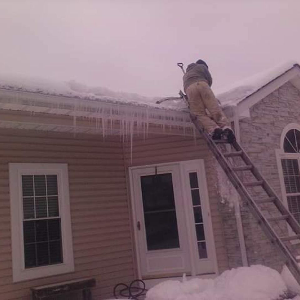 Worker on a ladder removing ice and snow buildup from a home's roof and gutters.