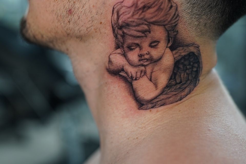 Image of a cherub angel with eyes closed and arms folded located on the customer neck left side hugging his hairline
