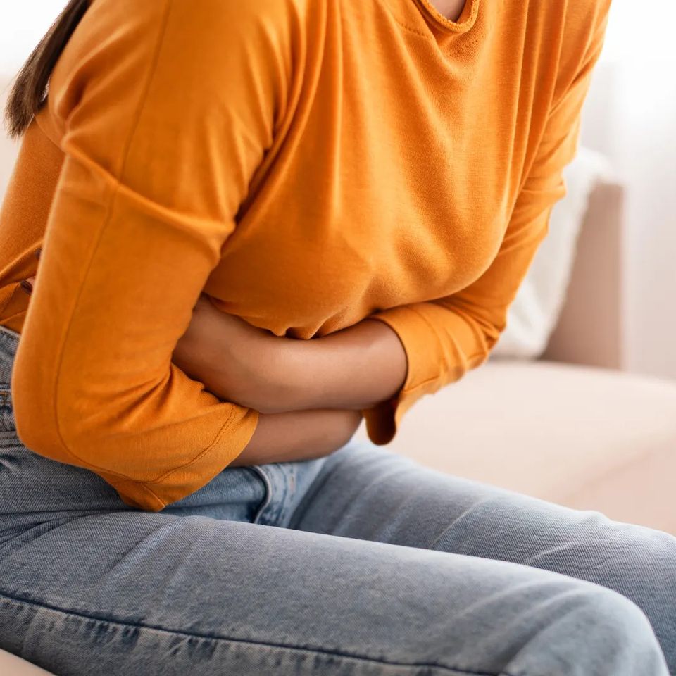 Woman in an orange top holding her stomach on a couch seeking Alleviate PMS cramp relief IV therapy in Palm Beach County FL