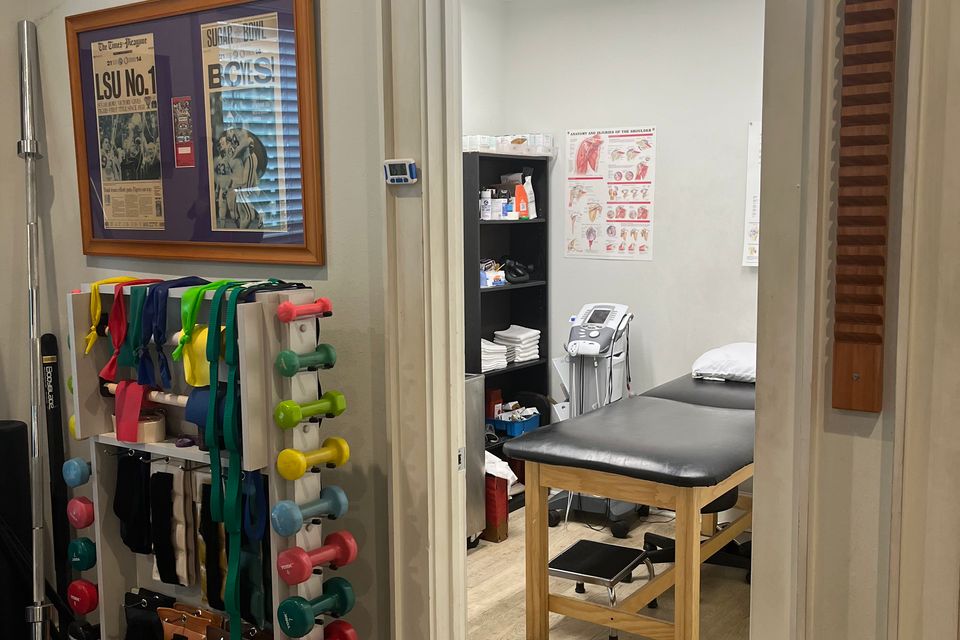 Interior view of a therapy room at Mid City Physical Therapy in New Orleans, featuring exercise equipment including a Valor wheel, medicine balls, dumbbells, resistance bands, and a volleyball on a rack near a window with blinds.