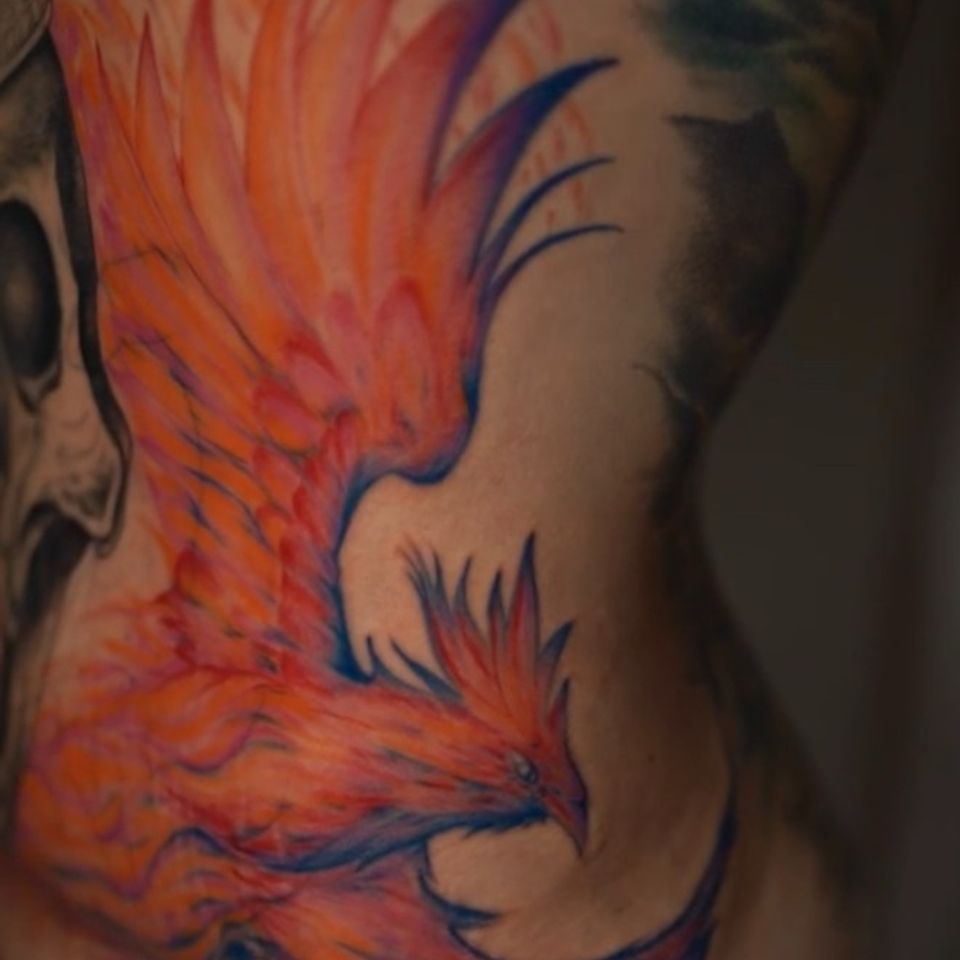 Image of a Tattoo on a womans left front side just under her breast extending down to her hip and a lot of skin in this area. Tattoo is bright orange, read and blue making the phoenix look like it's on fire