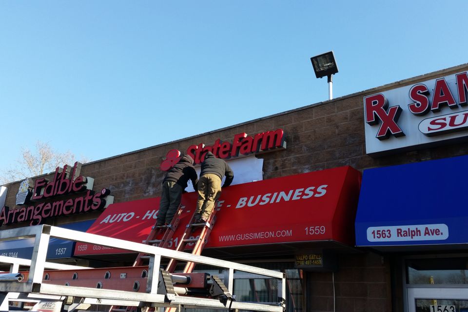 Two installers fitting a red printed storefront awning below dimensional channel letters on a Brooklyn commercial strip