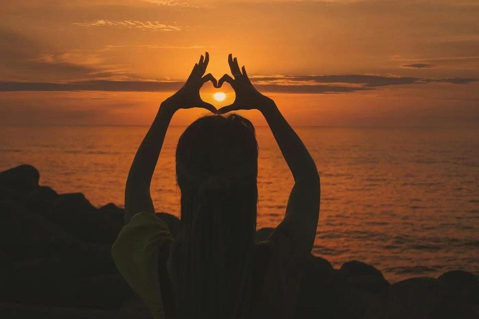 Silhouette of a person making a shape of a heart with their hands around the sun over a body of water at sunset 