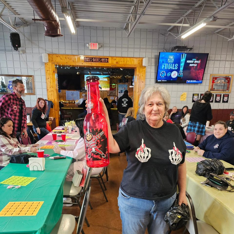 Woman holding a large red bottle prize at a community bingo event with participants seated at tables in the background.