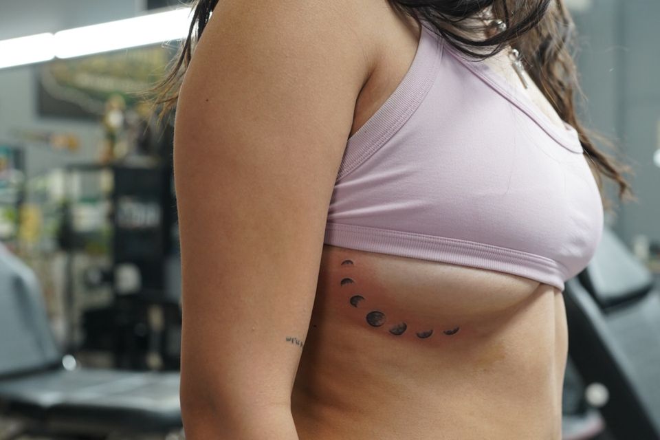 Image of a tattoo under a woman's right breast from a sliver to a full moon and variations from sliver to full in black ink