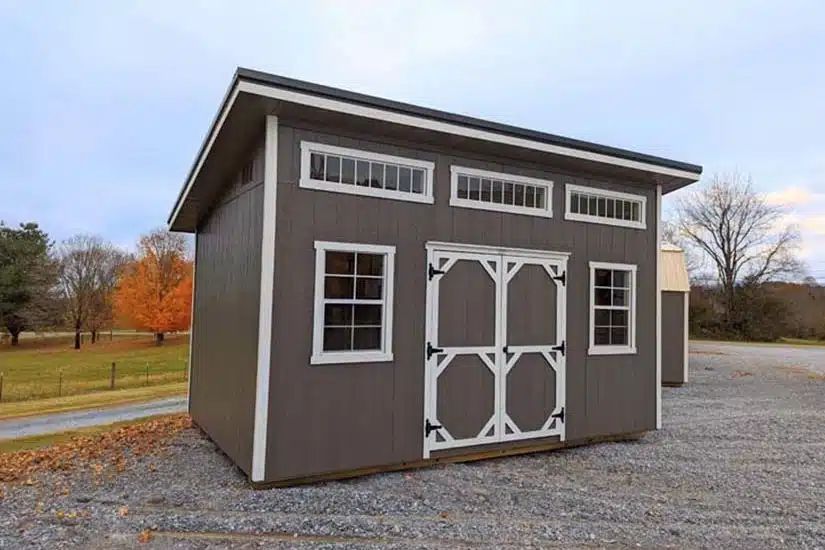 Gray studio shed with transom windows and white double doors on a gravel lot in East Tennessee