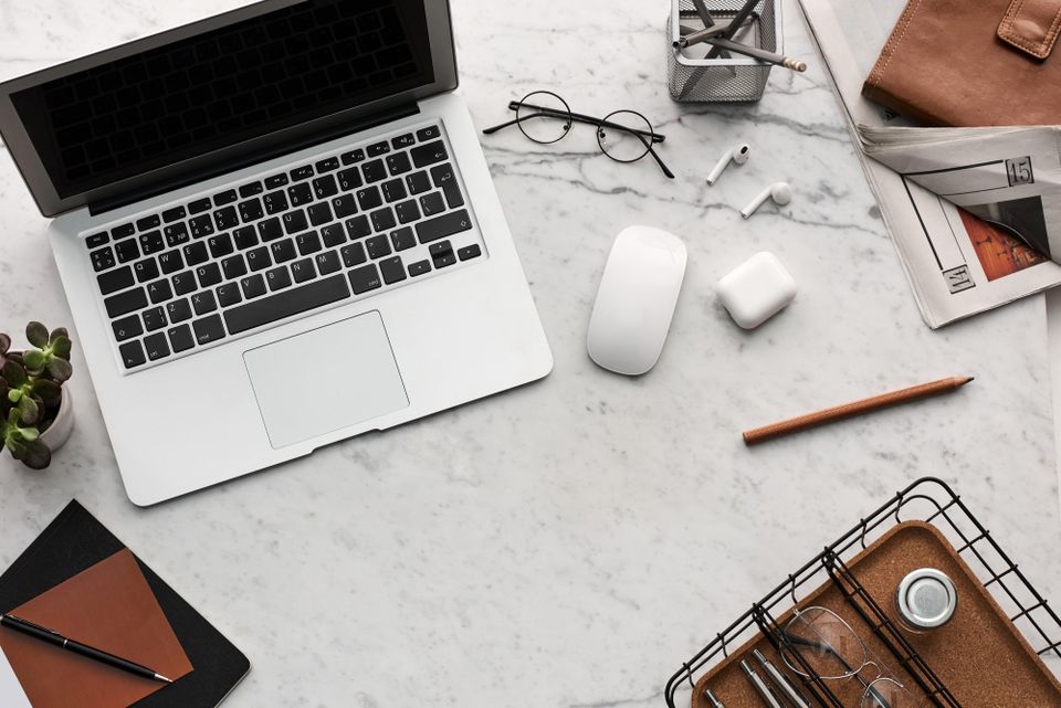 Desk with laptop, a wallet, some glasses and writing tools seen from above.