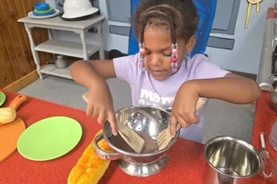 A child is mixing in a metal bowl using imagination, at a child's table with fake bread.