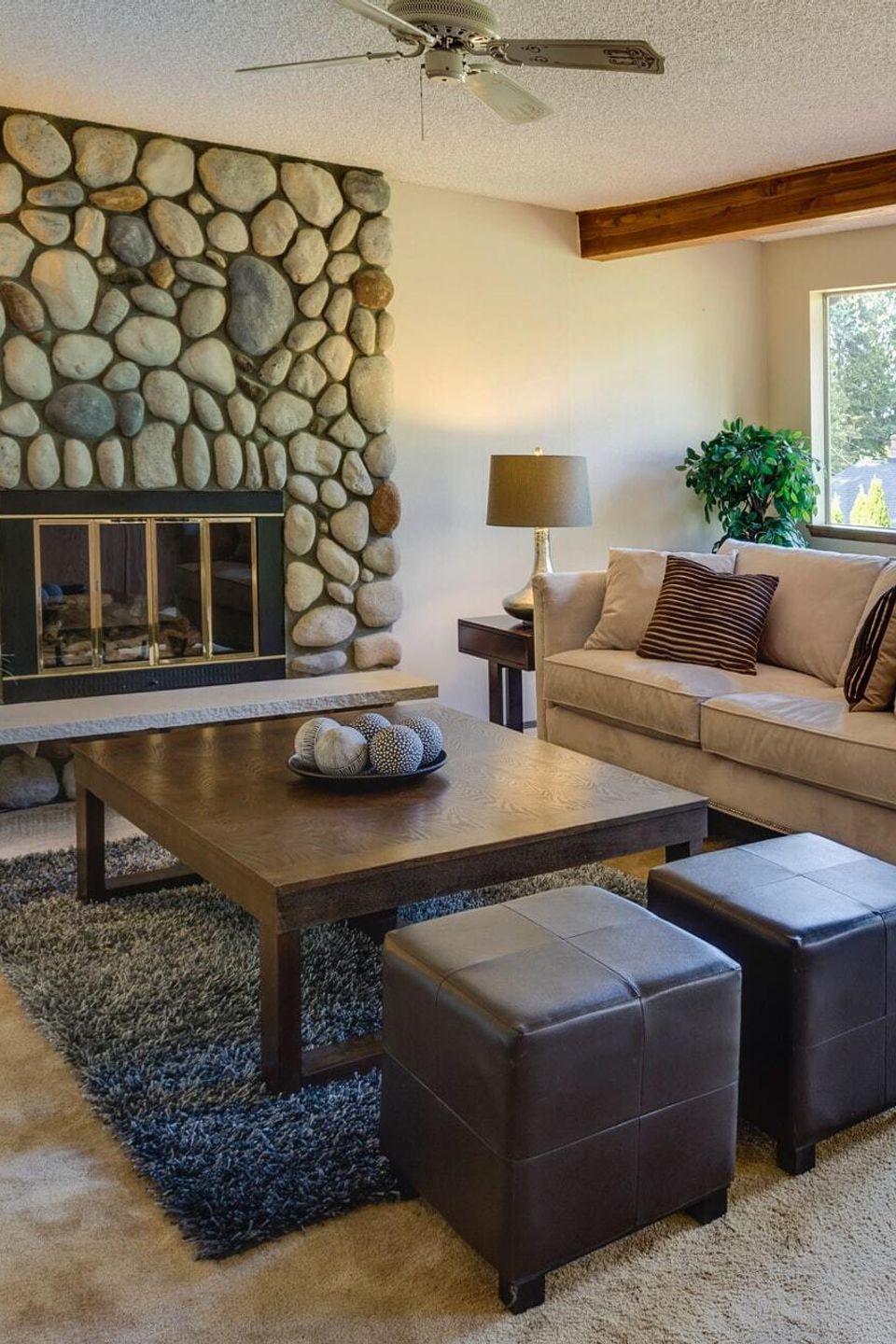 Cozy living room with stone fireplace, beige sofa, wooden coffee table, leather ottomans, and large windows overlooking deck and greenery