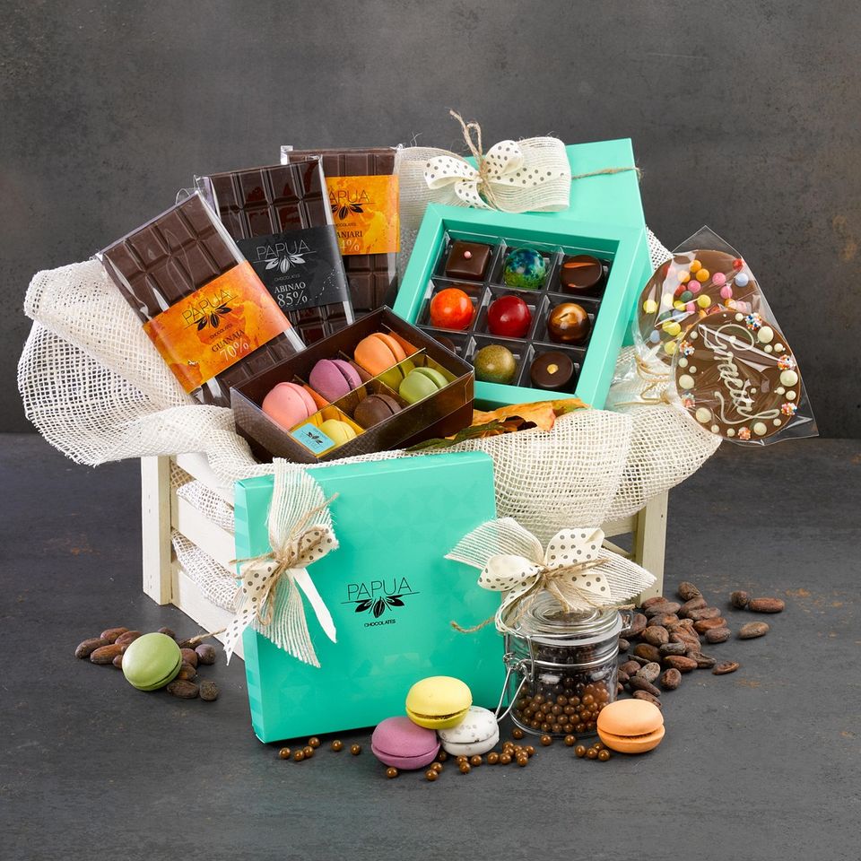 Gift basket with artisan chocolates, colorful macarons, a heart-shaped chocolate lollipop, coffee beans, and a turquoise gift bag, arranged with burlap and mesh fabric. Chocolate baskets