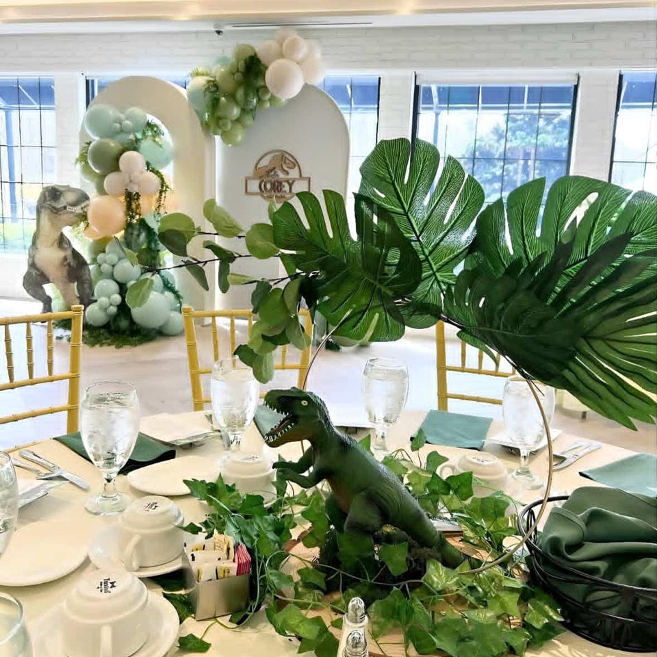 urassic-themed party with dinosaur table centerpiece, tropical greenery, balloon arch, and safari-style decor backdrop