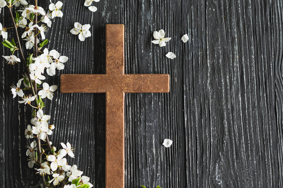 Wooden cross resting on a dark wood background surrounded by delicate white flowers, symbolizing faith, hope, and spiritual devotion.