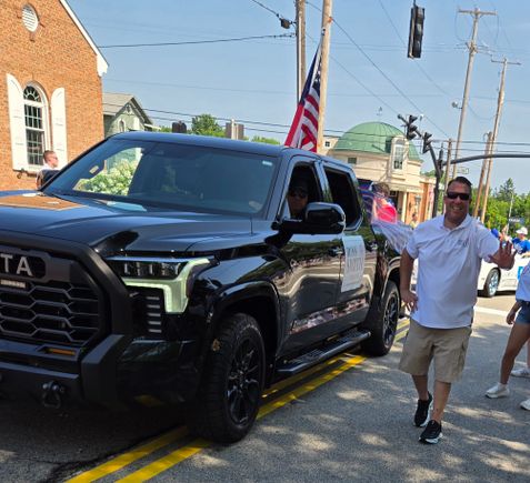 Man waving beside a black truck decorated with an American flag during the Fourth of July parade.