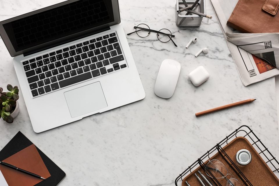 Desk with laptop, a wallet, some glasses and writing tools seen from above.