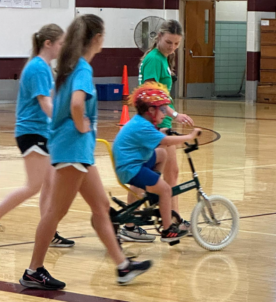 A volunteer coach walks alongside a child riding an adaptive bicycle while other coaches observe during DSAV's iCan Shine Bike Camp.