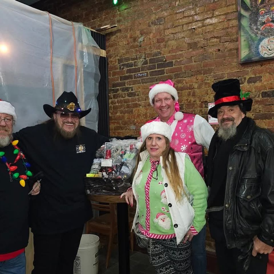 Group of attendees in holiday attire posing indoors beside raffle baskets during a Christmas-themed gathering.