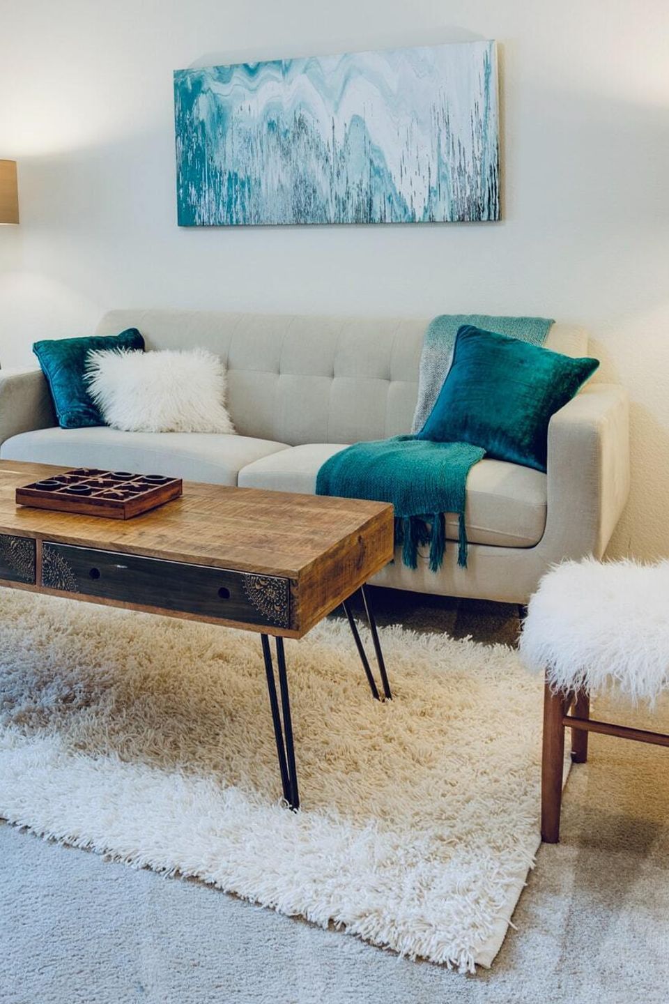 Modern living room with beige tufted sofa, teal pillows, rustic wooden coffee table, fluffy stools, and abstract teal artwork