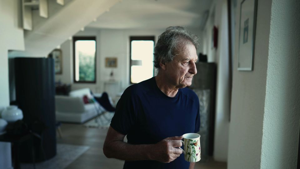 A contemplative mature man stands at home drinking cup of coffee or tea at living room.
