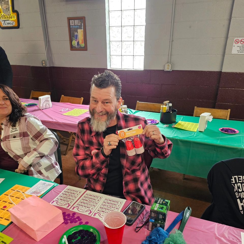 Man seated at a bingo table holding a package of mini cups as a prize, surrounded by bingo cards, markers, and event decorations.