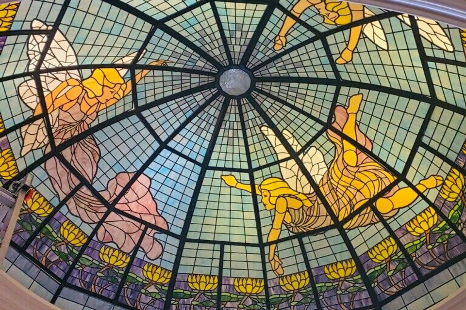 An upward view of a large, circular stained-glass ceiling dome featuring yellow figures and floral patterns, illuminated by professionally installed accent lighting.