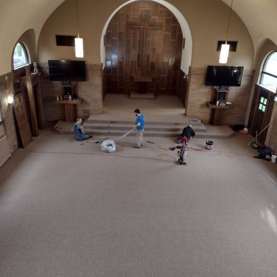 Workers installing carpet in a church sanctuary with wood doors, steps, and arched windows.