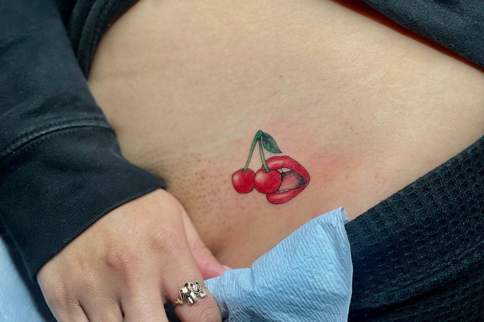 Image of a woman's pulling down her pants to show a colorful red and green and black ink tattoo of a mouth biting into a cherry located by her stomach just above her private parts