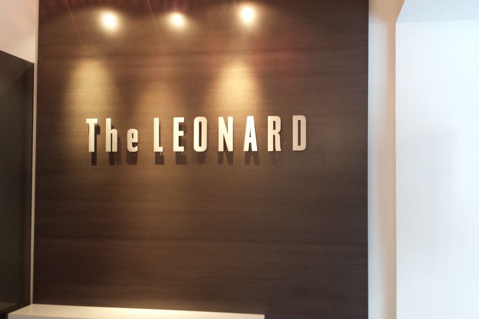 Flat cut aluminum letters reading The Leonard mounted on a dark wood panel wall under spotlights