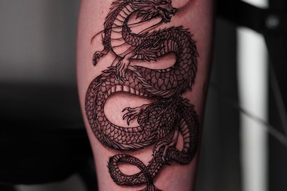 Image of a very detailed dragon tattoo on a man's right outside calf with a sun in the background in black and a hint of red/orange
