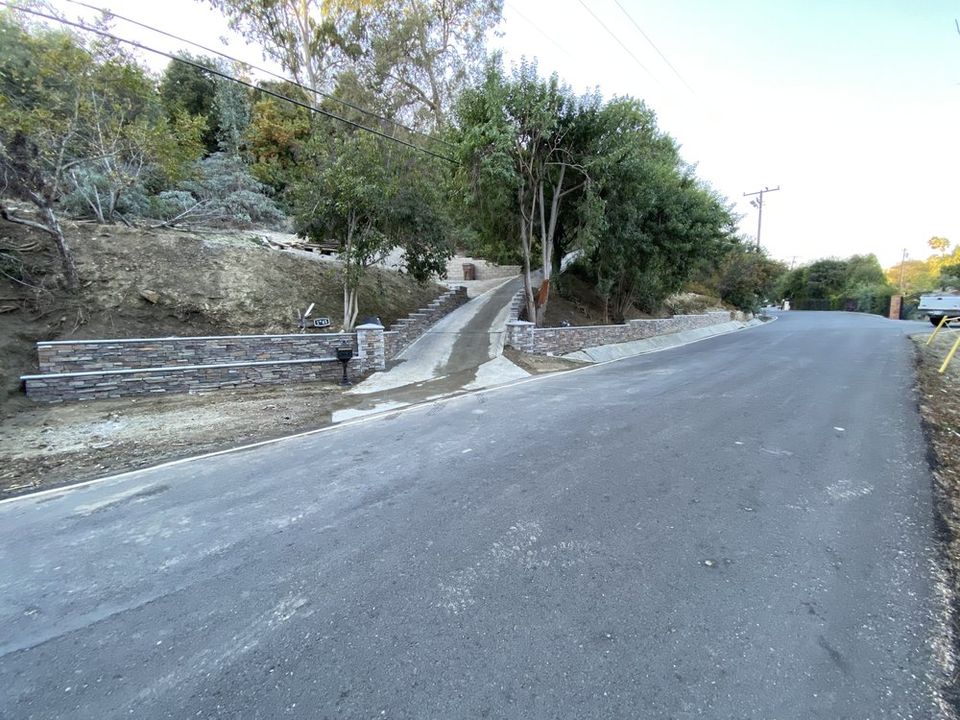 Wide concrete driveway leading to hillside property with decorative retaining wall and landscaped surroundings