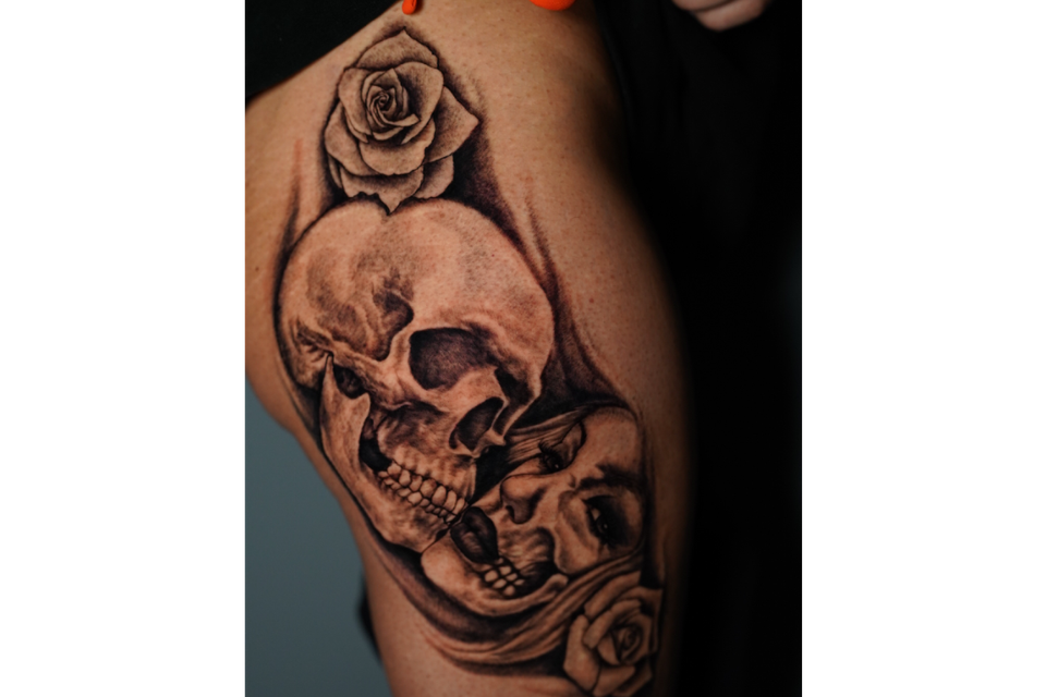 Image of a tattoo of a full skull and womans face with a her teeth and check exposed as if she is becomeing a skeleton with two flowers one on each side in black ink