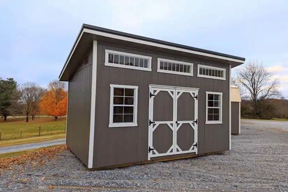 Gray studio shed with transom windows and white double doors on a gravel lot in East Tennessee