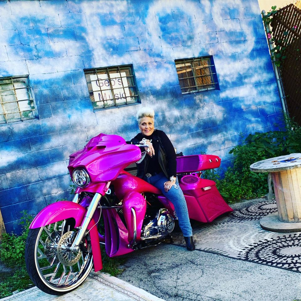 Kelly sitting on a custom pink touring motorcycle in front of a blue-painted building with a colorful urban backdrop.