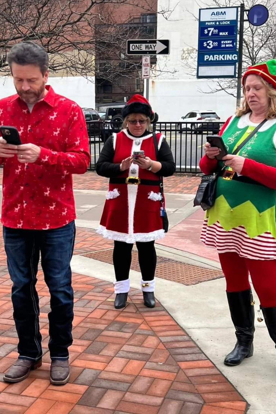 Three attendees dressed in Christmas-themed outfits standing on a downtown sidewalk while looking at their phones.