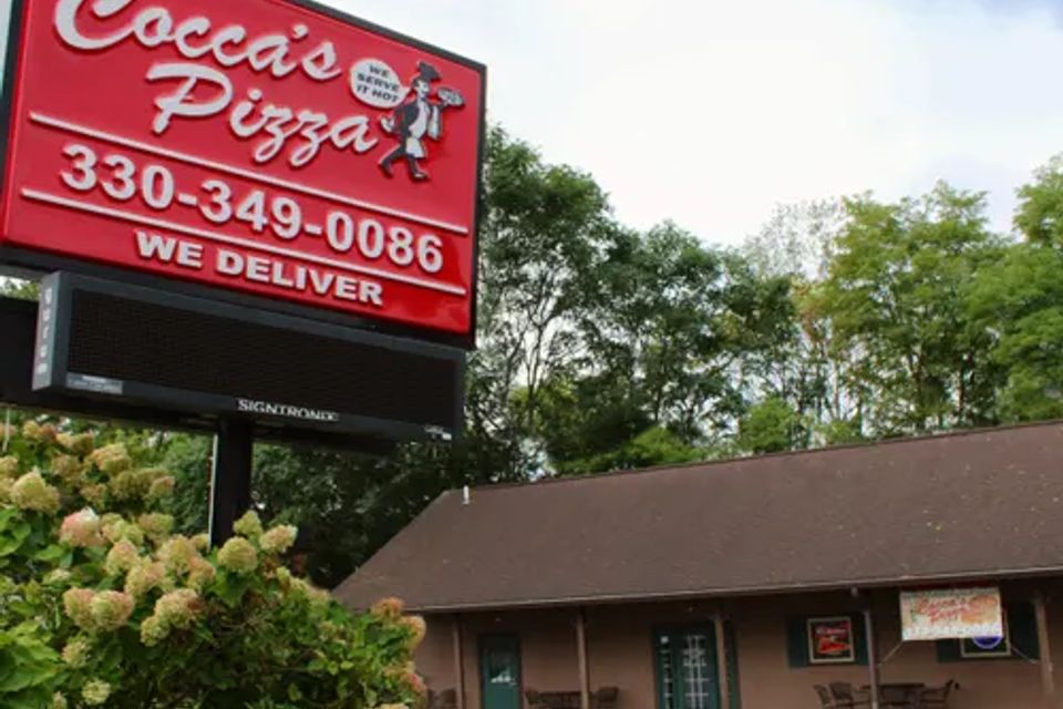 Cocca’s Pizza Mineral Ridge, Ohio location featuring a large roadside sign, parking area, and brown storefront building surrounded by trees.
