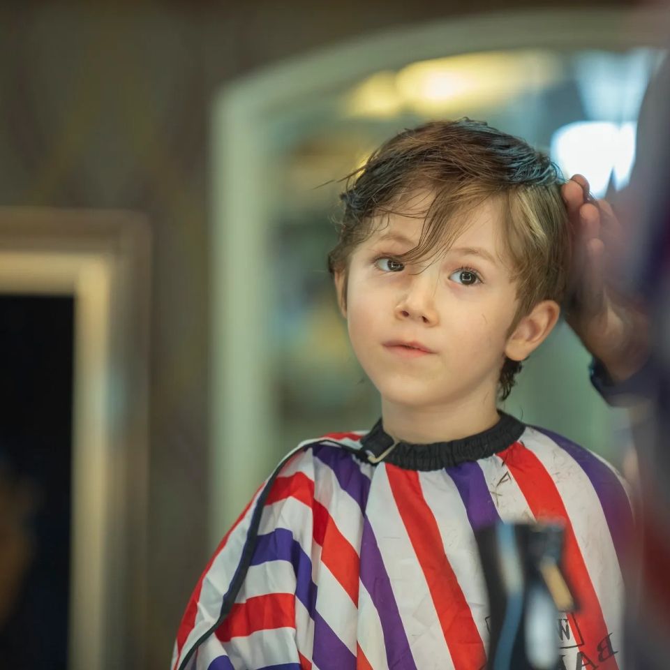 A close-up of a client receiving a haircut in a barbershop, with a barber adjusting their hair and a striped cape draped over them.