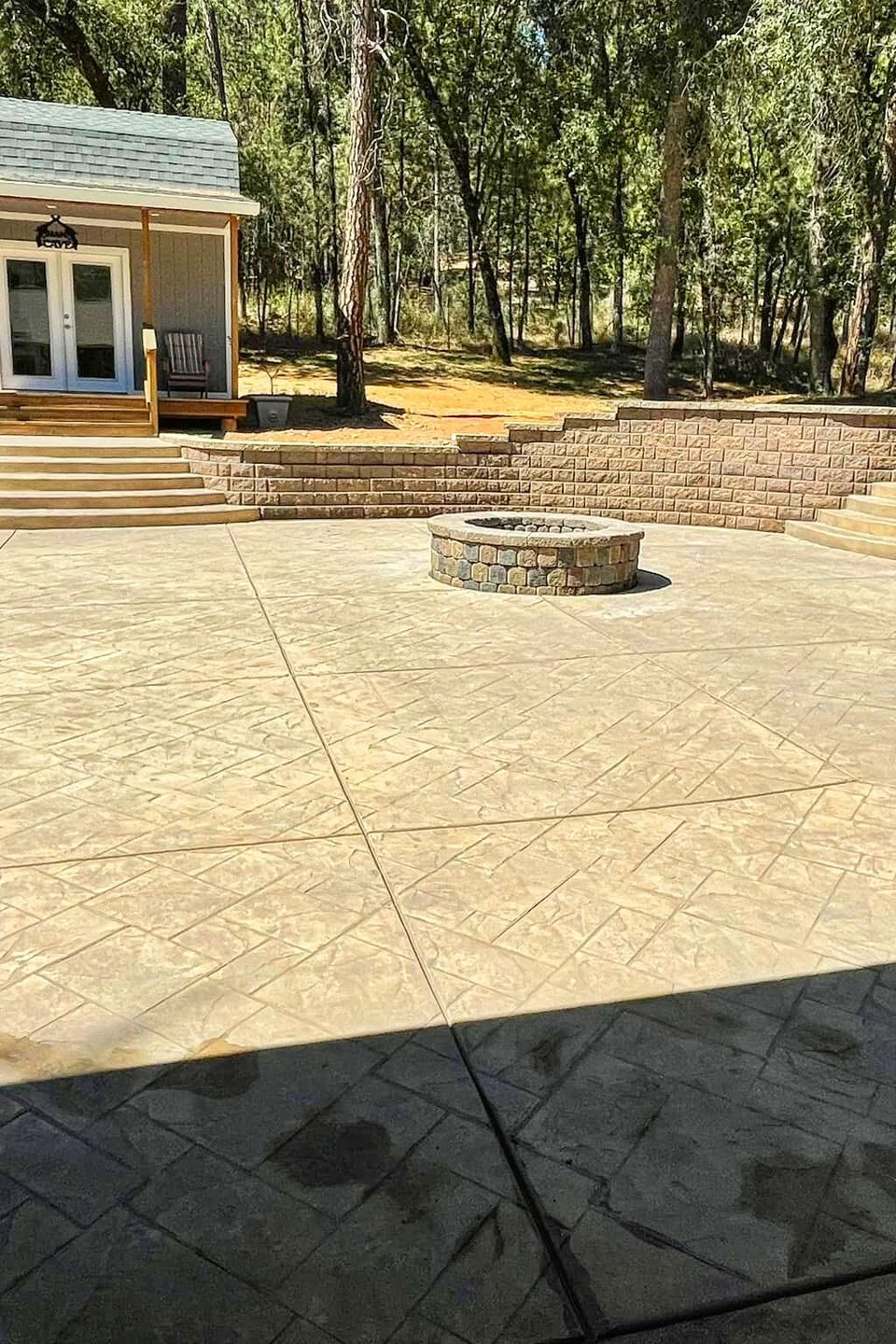 Completed backyard hardscape by Alta Vista Landscaping & Tree Service featuring a large stamped concrete patio, custom stone fire pit, and tiered retaining walls with integrated stairs and a modern garden cottage