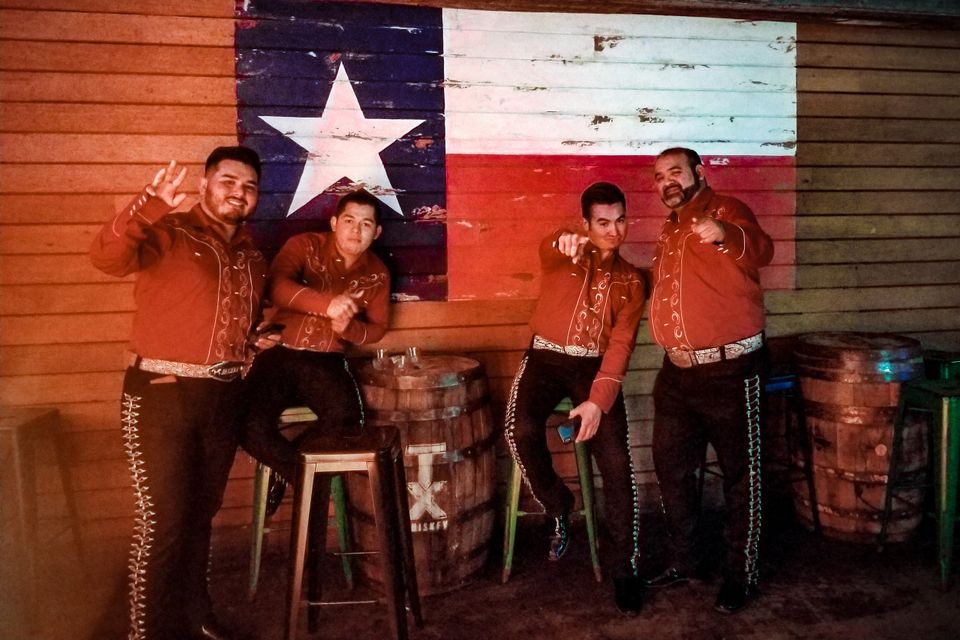 Four Mariachi Capital  musicians in red charro outfits posing in front of a rustic Texas flag mural inside a bar.