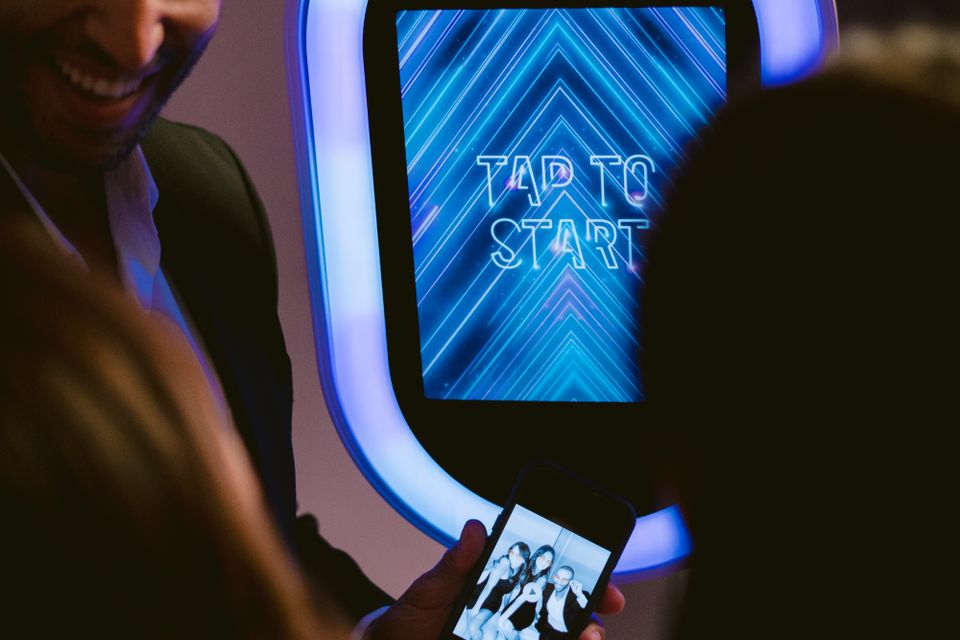 Guests standing by an A&M Photobooth with a “Tap to Start” screen while someone views a photo on their phone.